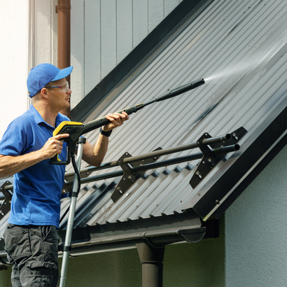 Man in blue shirt and cap power washing a corrugated metal roof.
