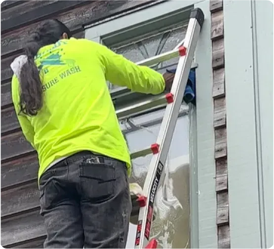 Person in neon green shirt washing a window with a ladder, outside a wooden building.