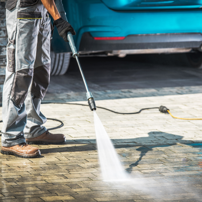 Person power washing a brick driveway near a teal car, water spraying.