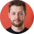A man with a beard and short brown hair, looking at the camera, stands in front of a red background.