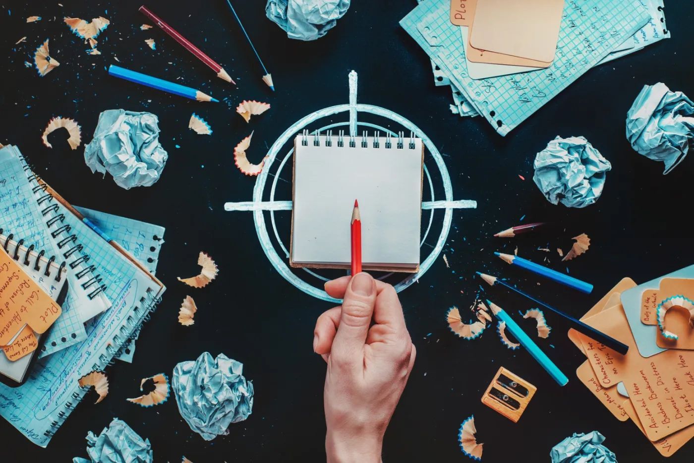 Hand holding a red pencil over an open notebook centered within a target.