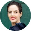 Woman with dark hair and red lipstick smiles outdoors, against a green door, wearing a dark blue top.
