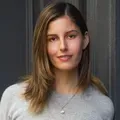 Woman with long brown hair wearing a grey sweater, standing in front of a grey background and smiling.