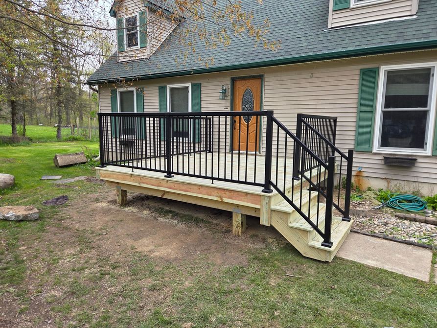 Small front porch with black railing and steps on a beige house, beside a grassy yard.