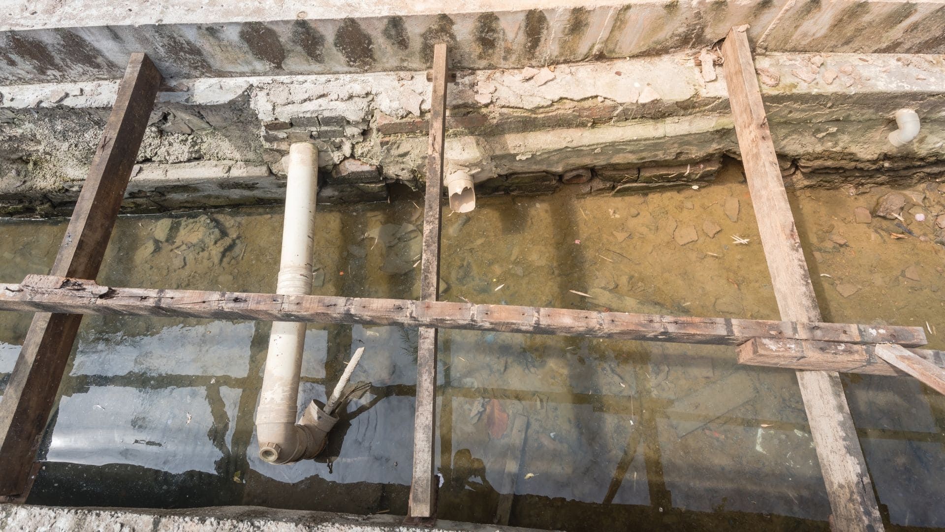 A construction site showing concrete trenches with wooden supports and exposed PVC pipes in muddy water.
