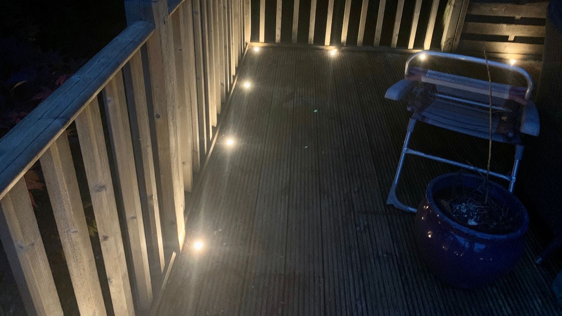 A wooden deck at night, illuminated by soft, recessed ground lights along the railing, with a chair and a plant pot.