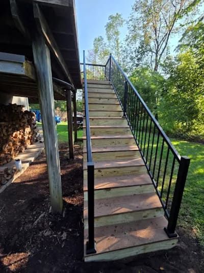 Wooden outdoor staircase with black metal railings leading up to a deck, set against a wooded background.