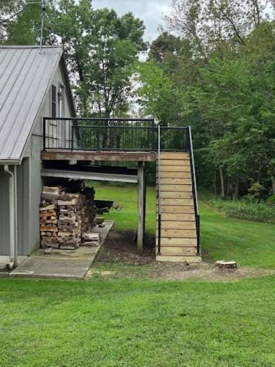 An elevated wooden deck with black railings and stairs stands beside a building with a stack of firewood underneath.