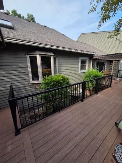 A wooden deck with black metal railings alongside a house with gray siding, two windows, and green shrubs.