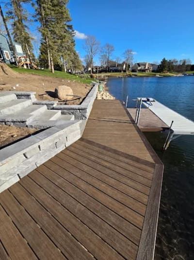 A lakefront property featuring a wooden deck with stone retaining walls and steps, leading to a metal dock on the water.