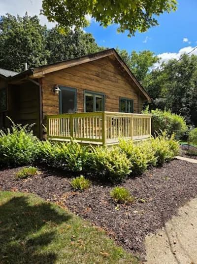 A log cabin exterior with a wooden deck, surrounded by green shrubs and mulch under a bright blue sky.