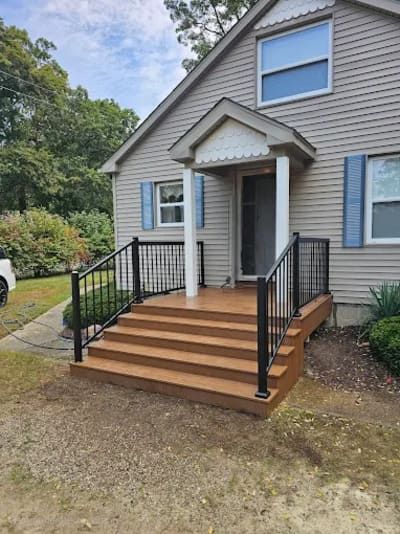 A newly installed wooden front porch with black railings and stairs leading to the entrance of a grey siding house.