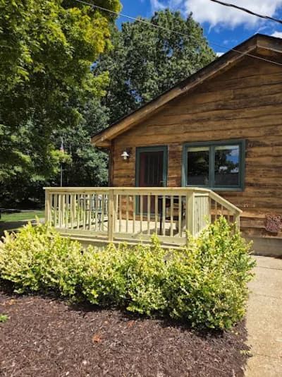 A log cabin with a light-colored wooden deck, surrounded by shrubs and trees under a partly cloudy blue sky.