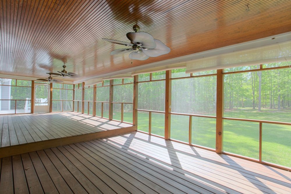 A screened-in porch with wood flooring and a paneled ceiling, featuring two ceiling fans and views of a green lawn.
