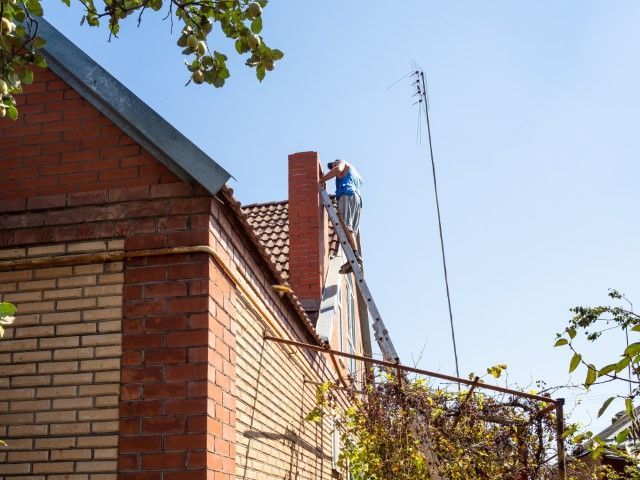 Worker on a roof beside a brick building, using a ladder under a clear blue sky.