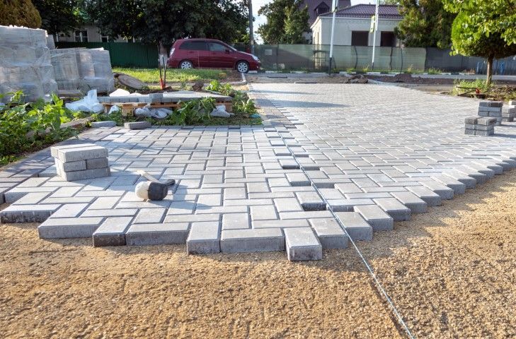 Paved walkway under construction with gray pavers, scattered bricks, and a red car in the background