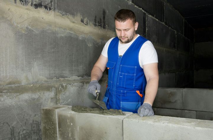 Worker in blue overalls stacking concrete blocks in an unfinished wall.