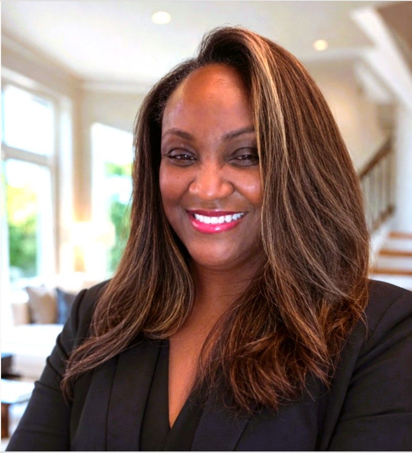 Smiling woman in a black blazer standing indoors with a bright, blurred background