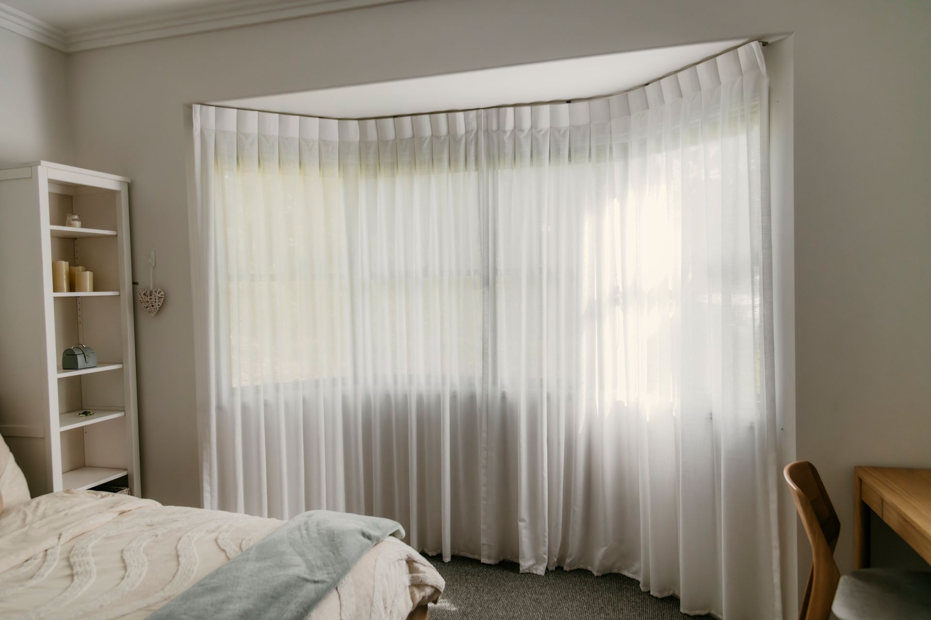 Bright bedroom with a curved window, sheer white curtains, bed, shelving, and a desk.