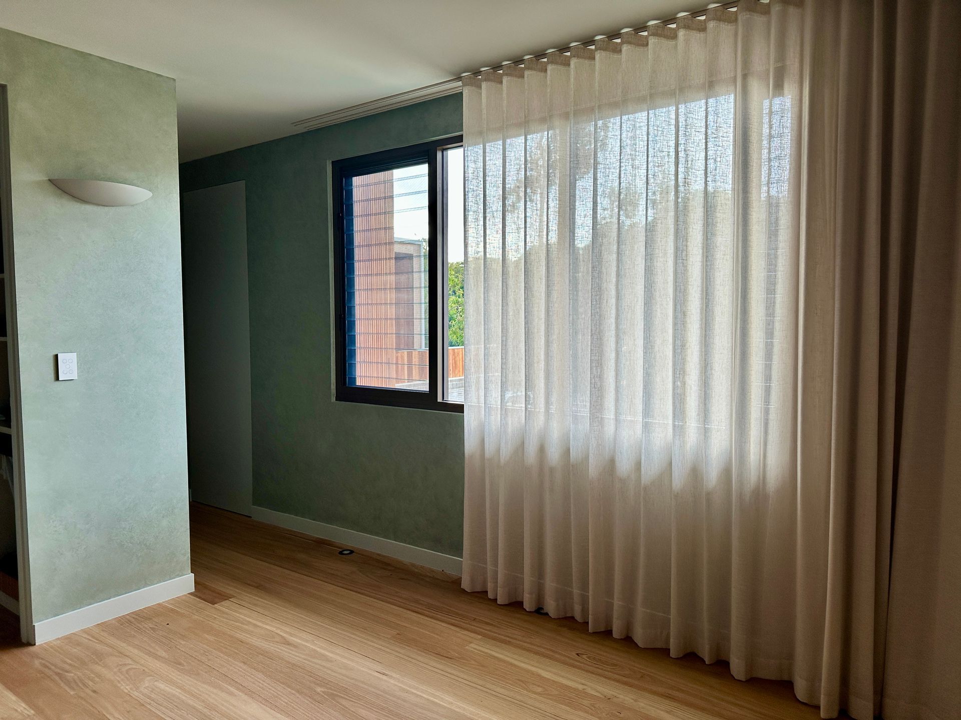 Empty room with green walls, wood floor, and a large window with sheer curtains.