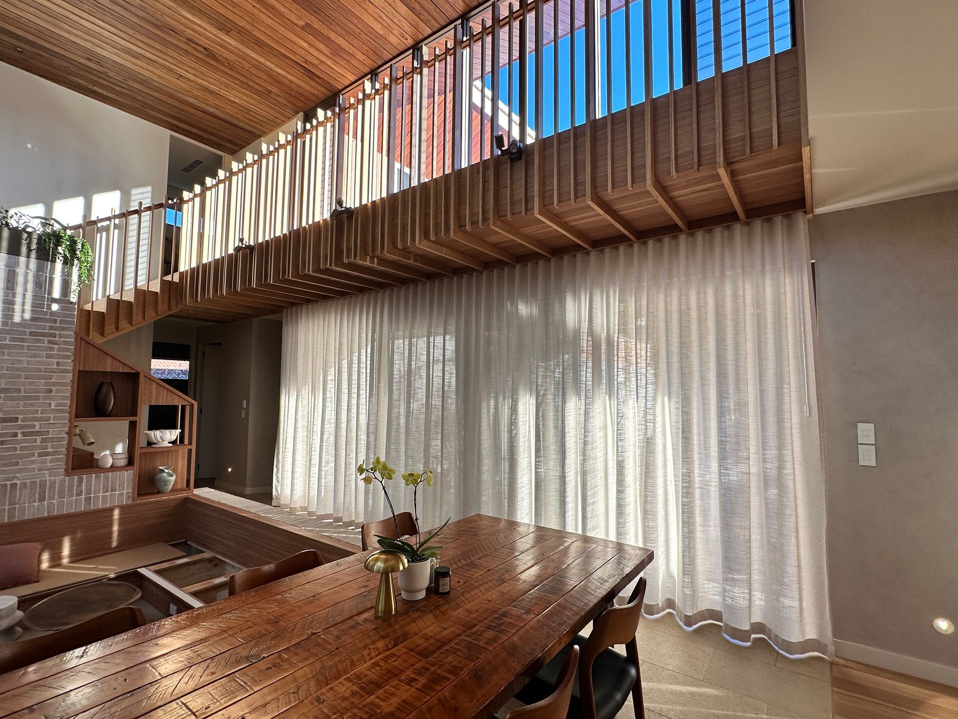 Modern dining room with wood ceiling, long table, and sheer white curtains beside a loft balcony
