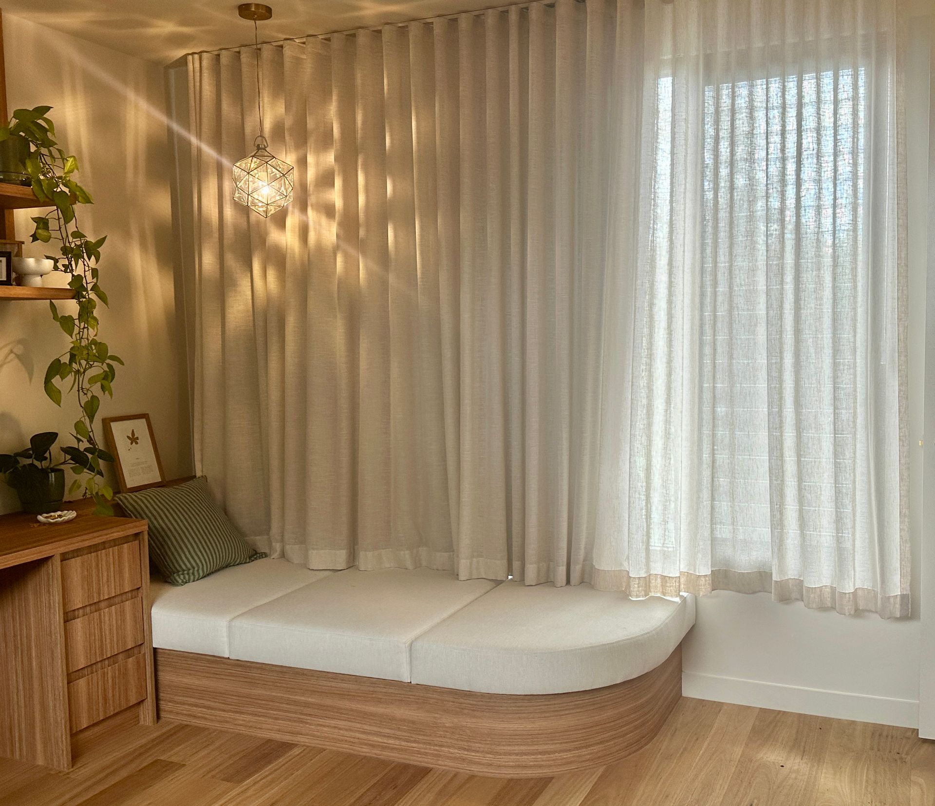 Cozy sunlit nook with sheer curtains, white cushioned bench, woven pendant light, and warm wood accents.