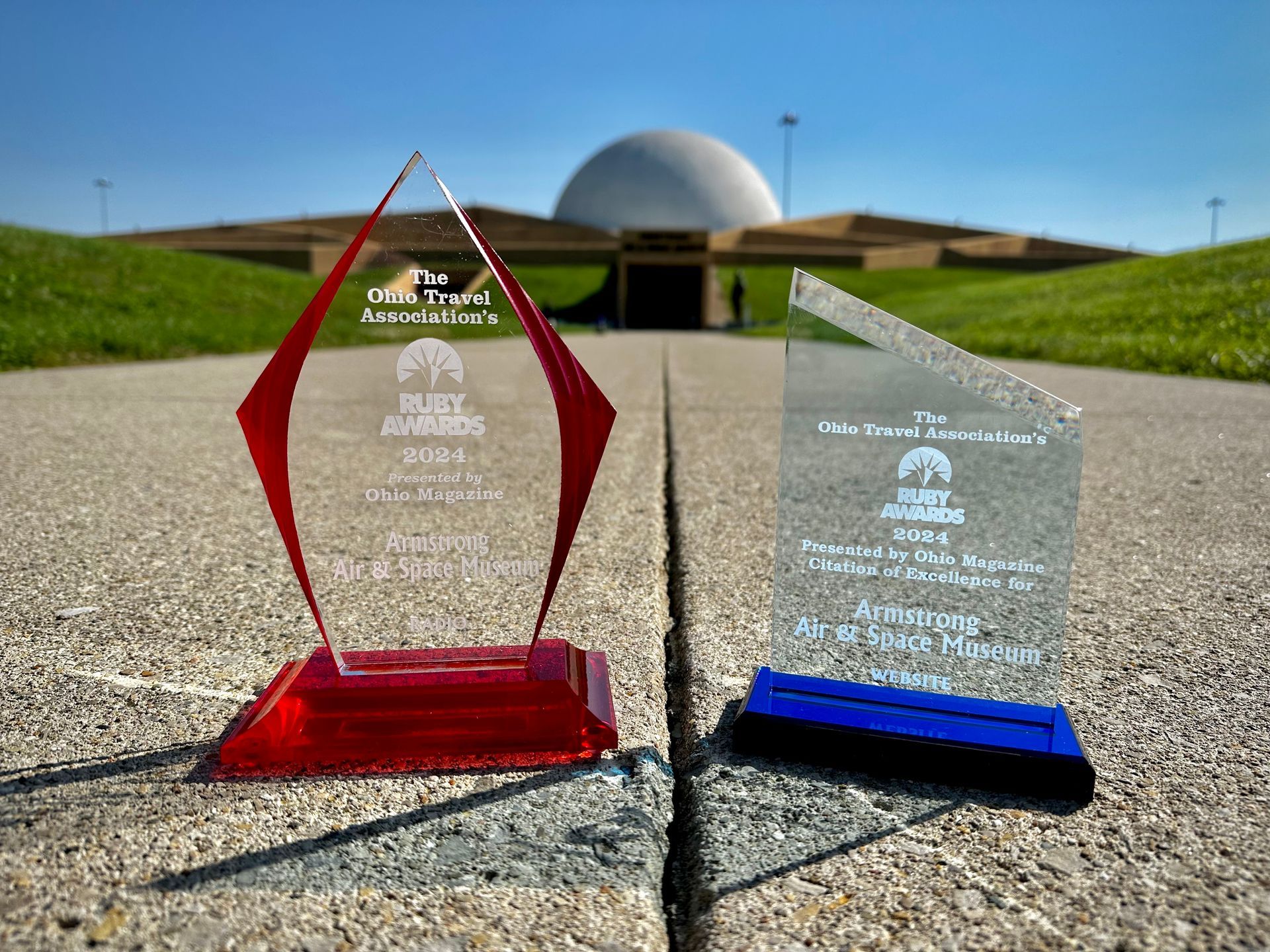 Two glass awards on a path with a dome building in the background. One award is red, the other is clear with a blue base.