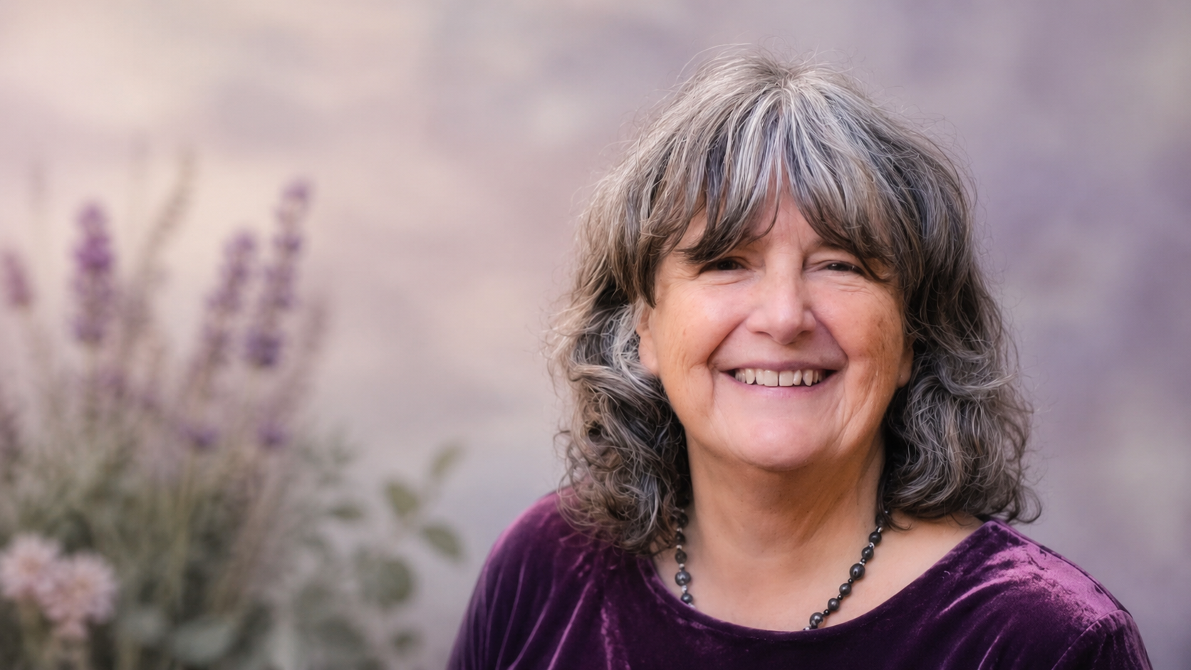 Smiling person with wavy gray hair in a purple top, standing outdoors with a soft floral background