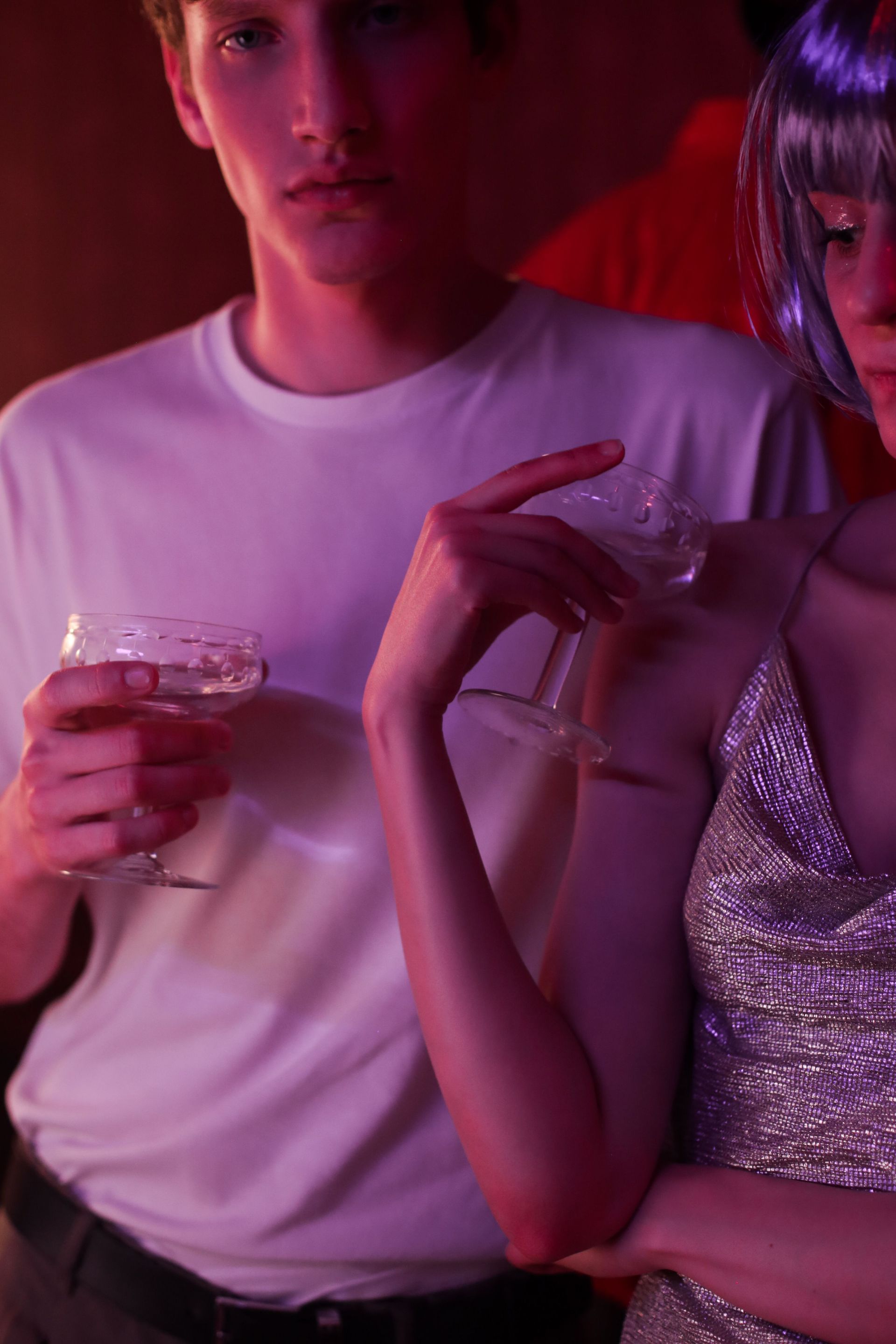 Two people at a nightclub under pink lighting, one holding a drink and the other in a silver dress reading a phone