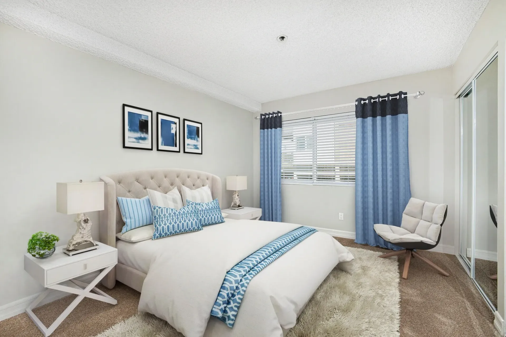Bright bedroom with a white bed, blue accents, bedside table, chair, and large window with curtains