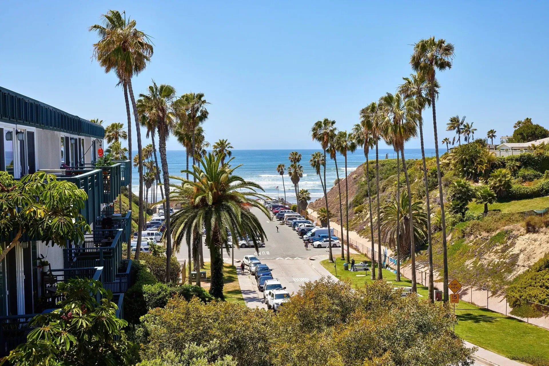 Palm-lined coastal path leading to a sunny beach and ocean view with distant pedestrians