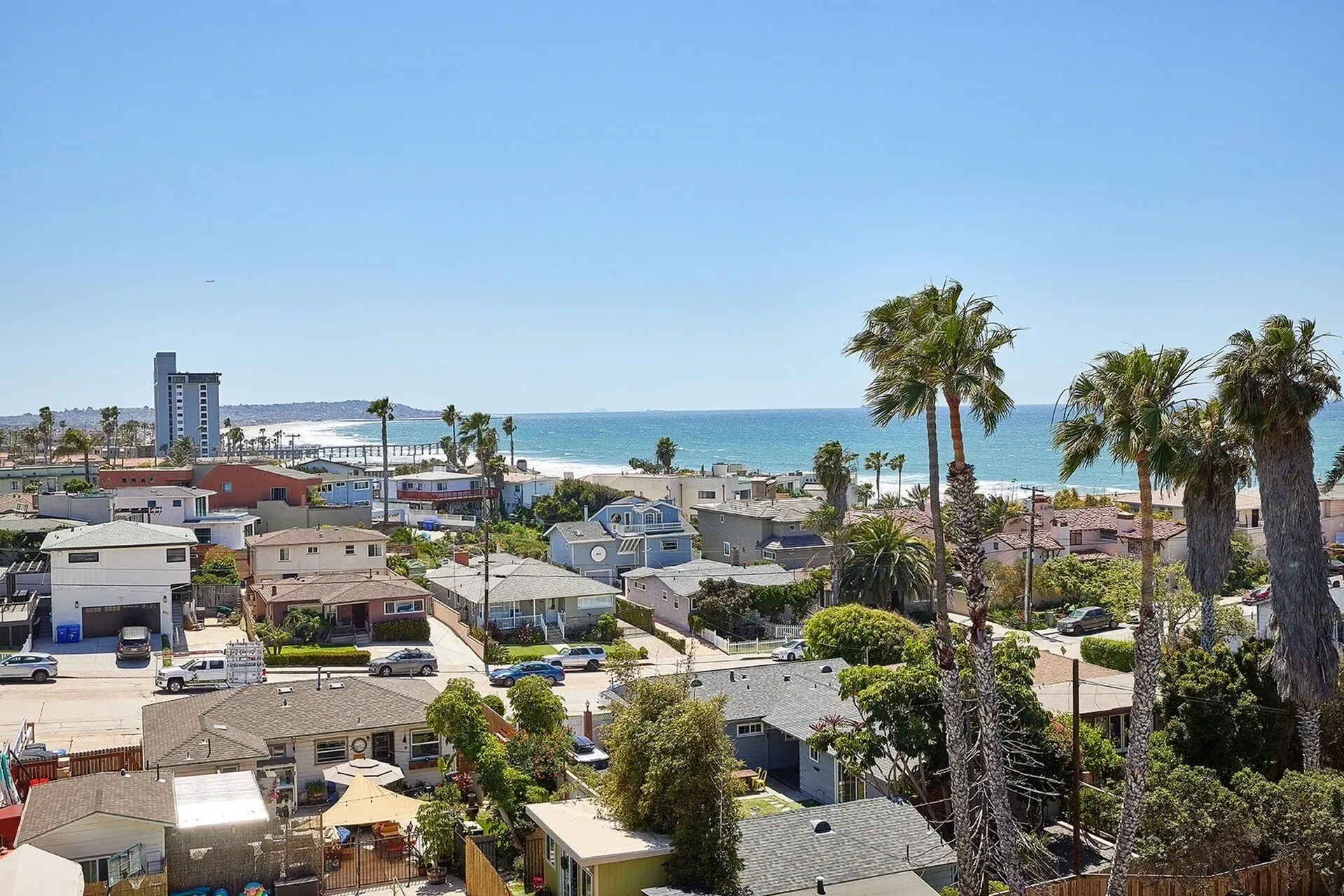 Coastal cityscape with palm trees, rooftops, and the ocean under a clear blue sky