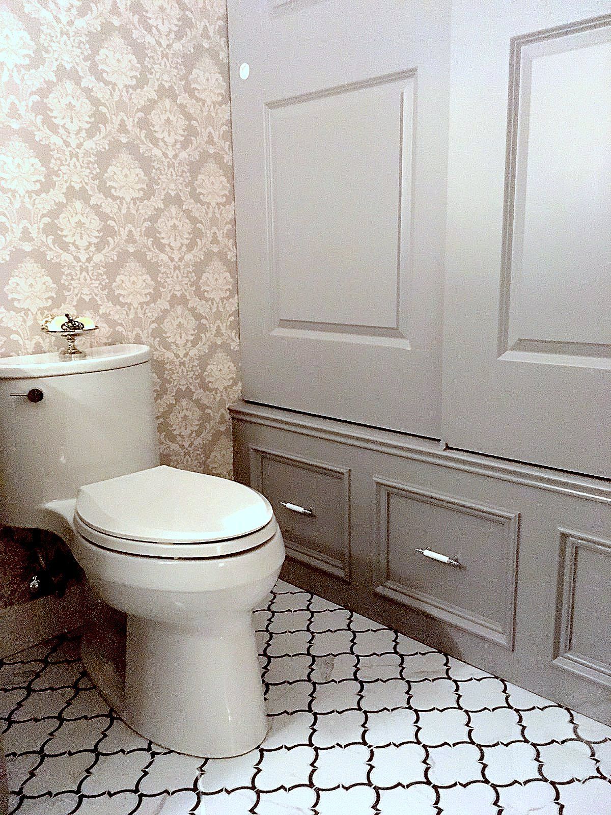 Small bathroom with a toilet, patterned wallpaper, gray cabinets, and white tile floor.