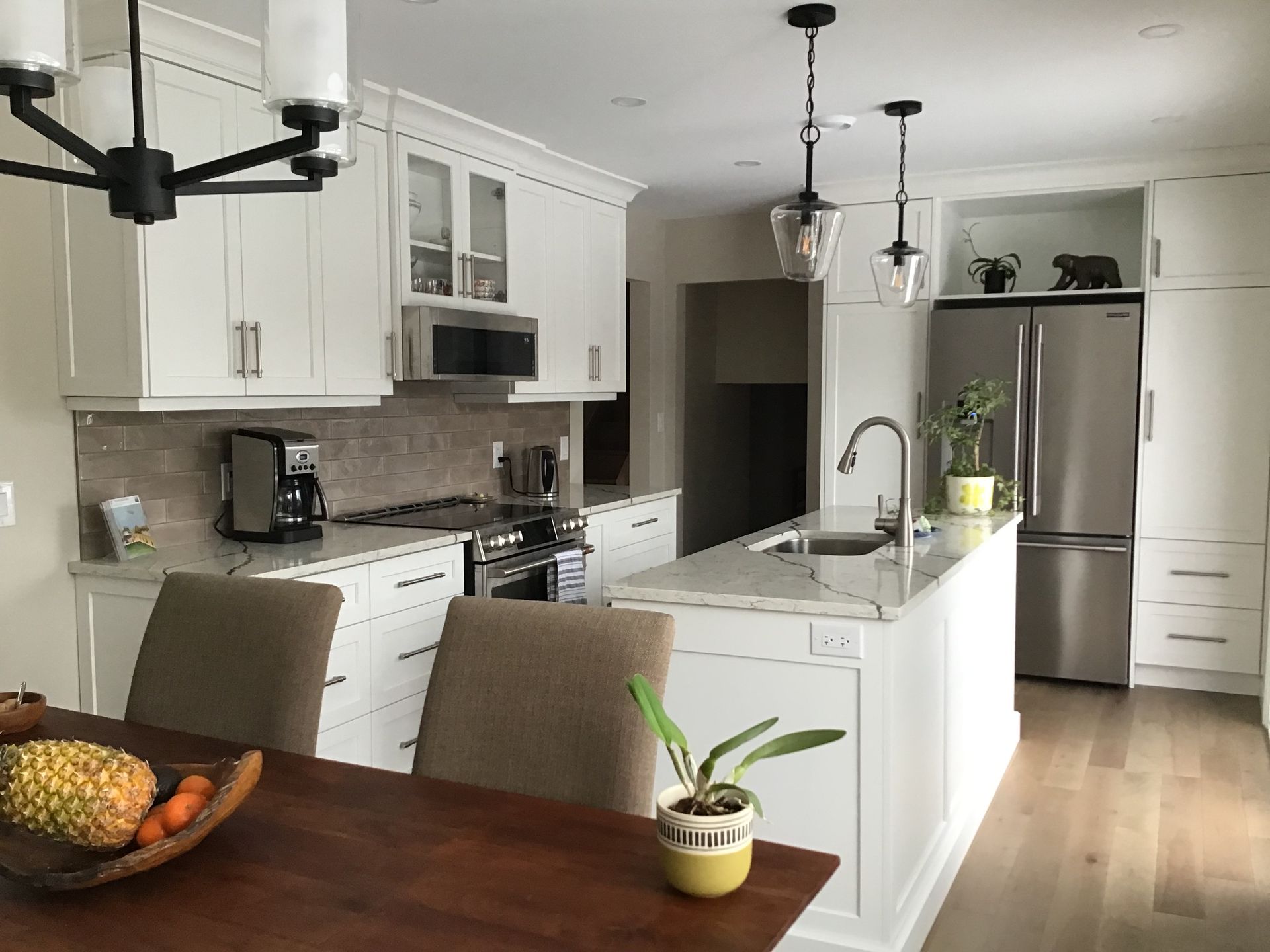 Bright white kitchen with island, stainless fridge, and wooden dining table with fruit bowl and small plant