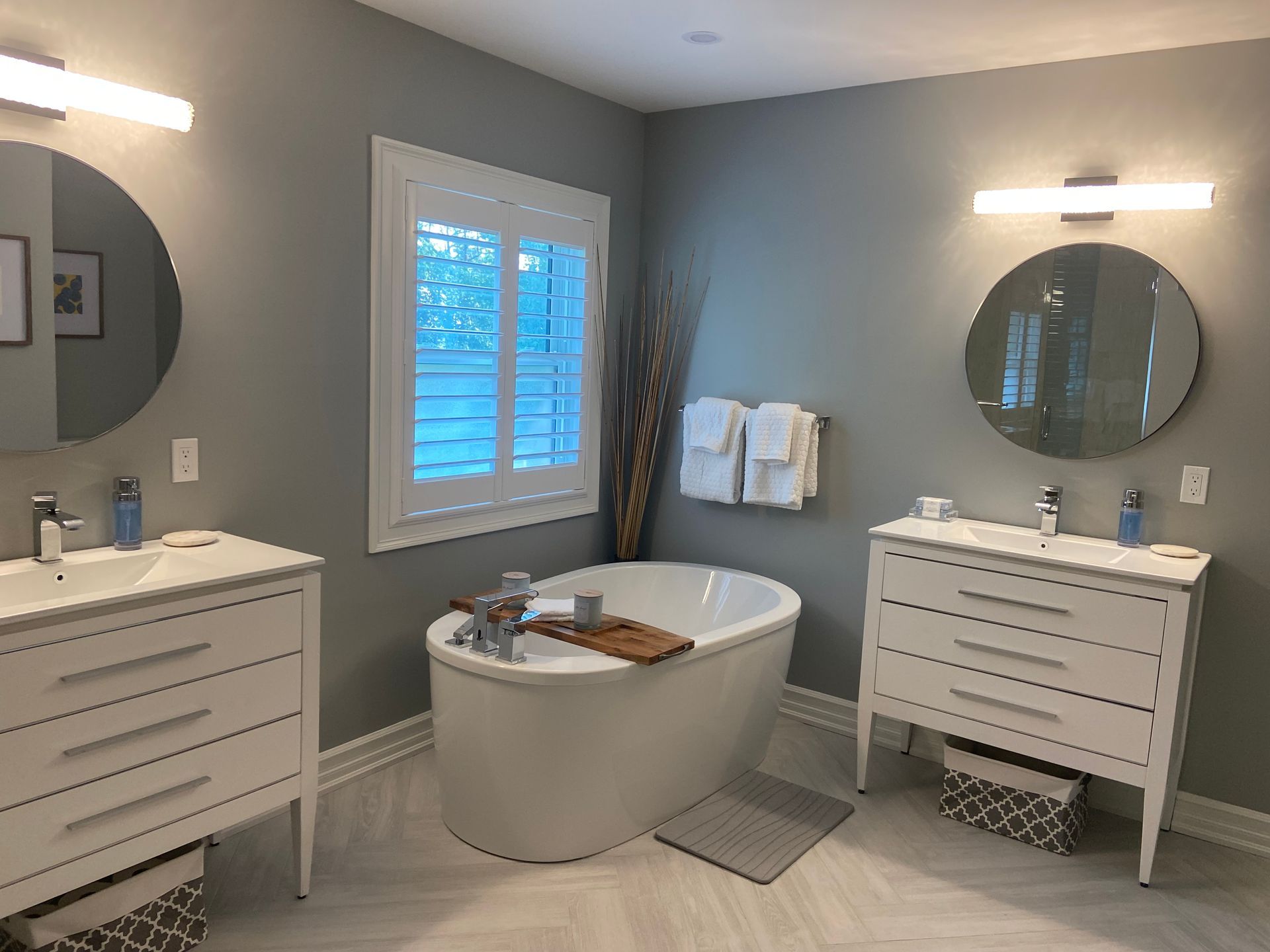 Bright modern bathroom with two white vanities, round mirrors, and a freestanding tub by a window