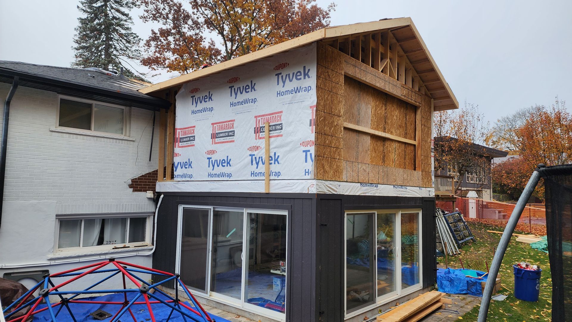House under construction with wrapped upper walls, wood framing, and a dark lower addition in a yard.