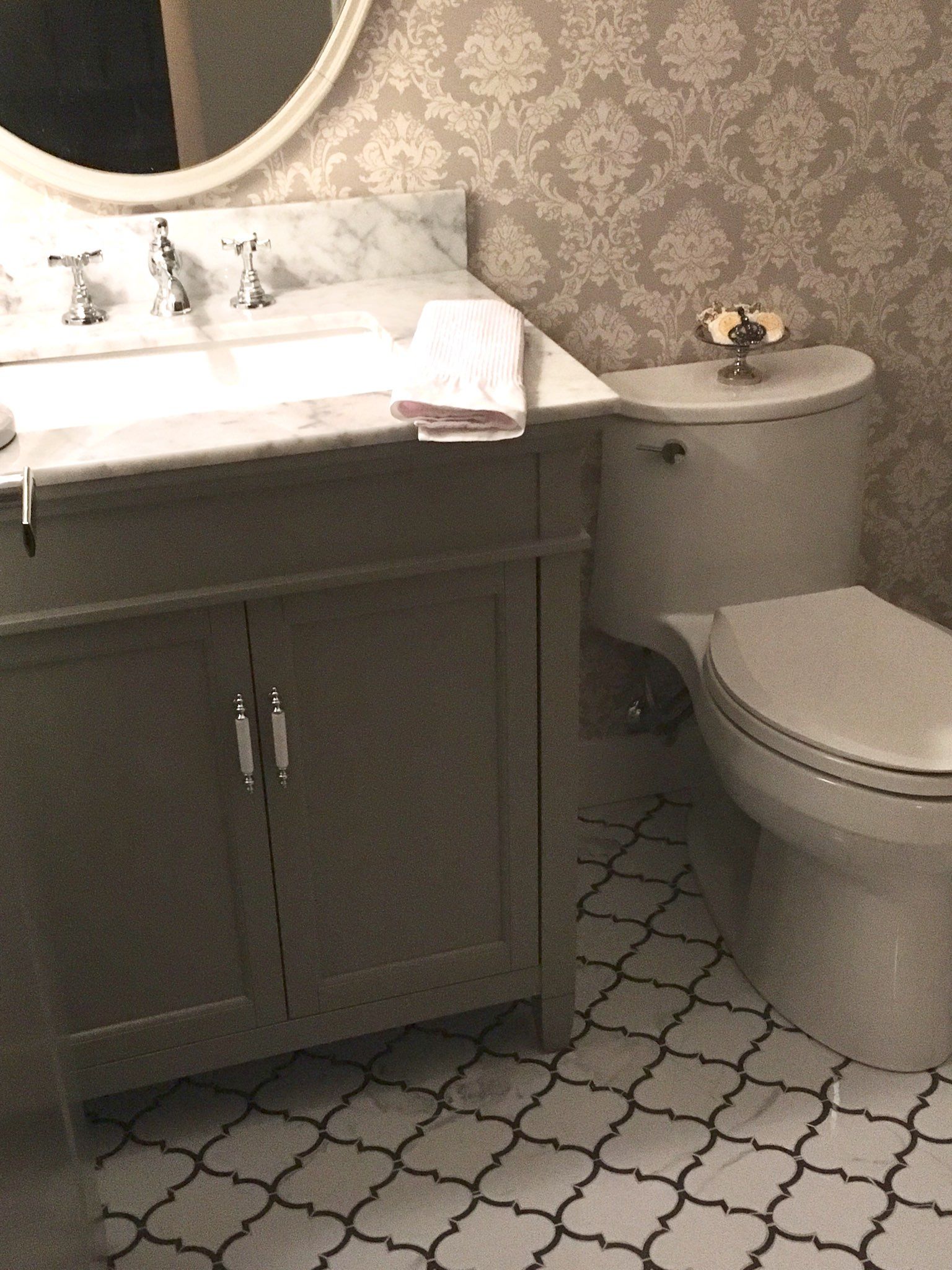 Small bathroom with gray vanity, round mirror, white toilet, and patterned tile floor.