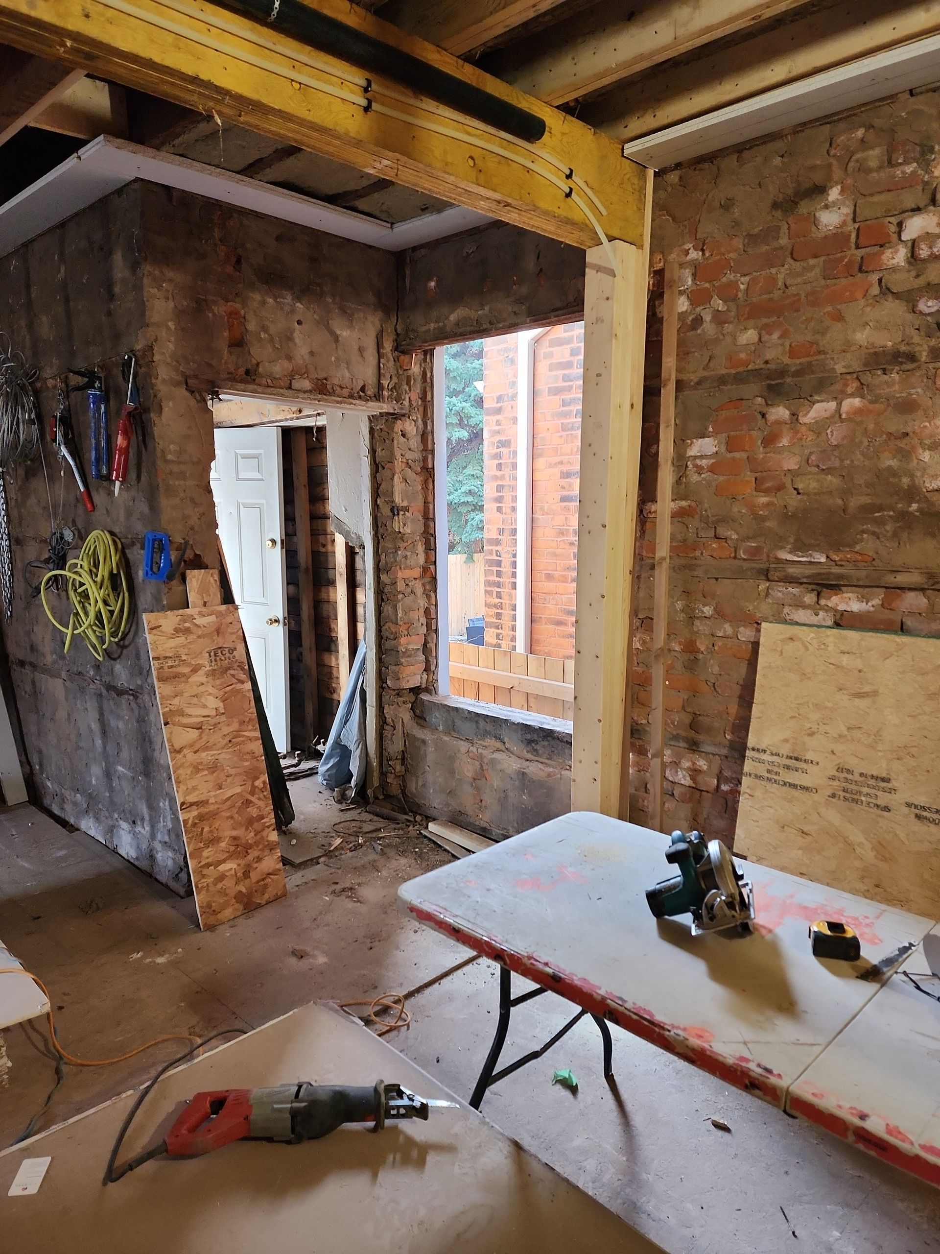 Renovation site with exposed brick walls, tools, and a partially built ceiling in a room with a window.