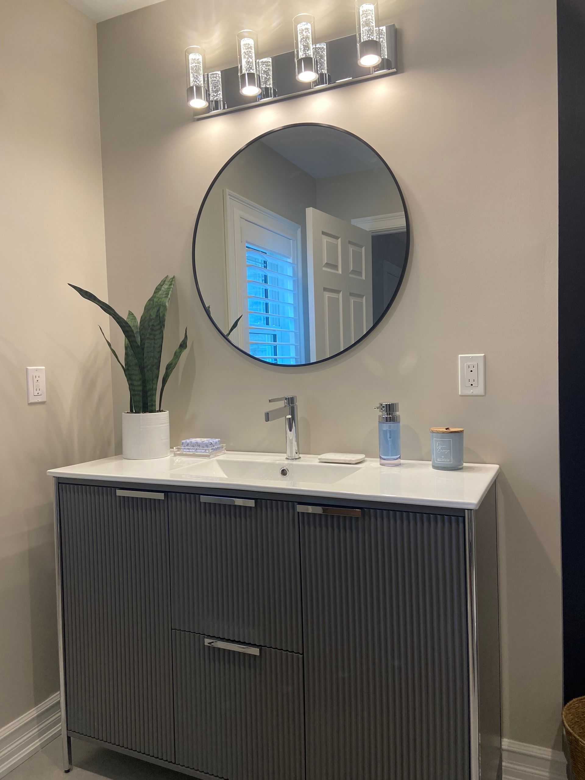 Modern bathroom vanity with round mirror, light fixture, plant, and countertop accessories