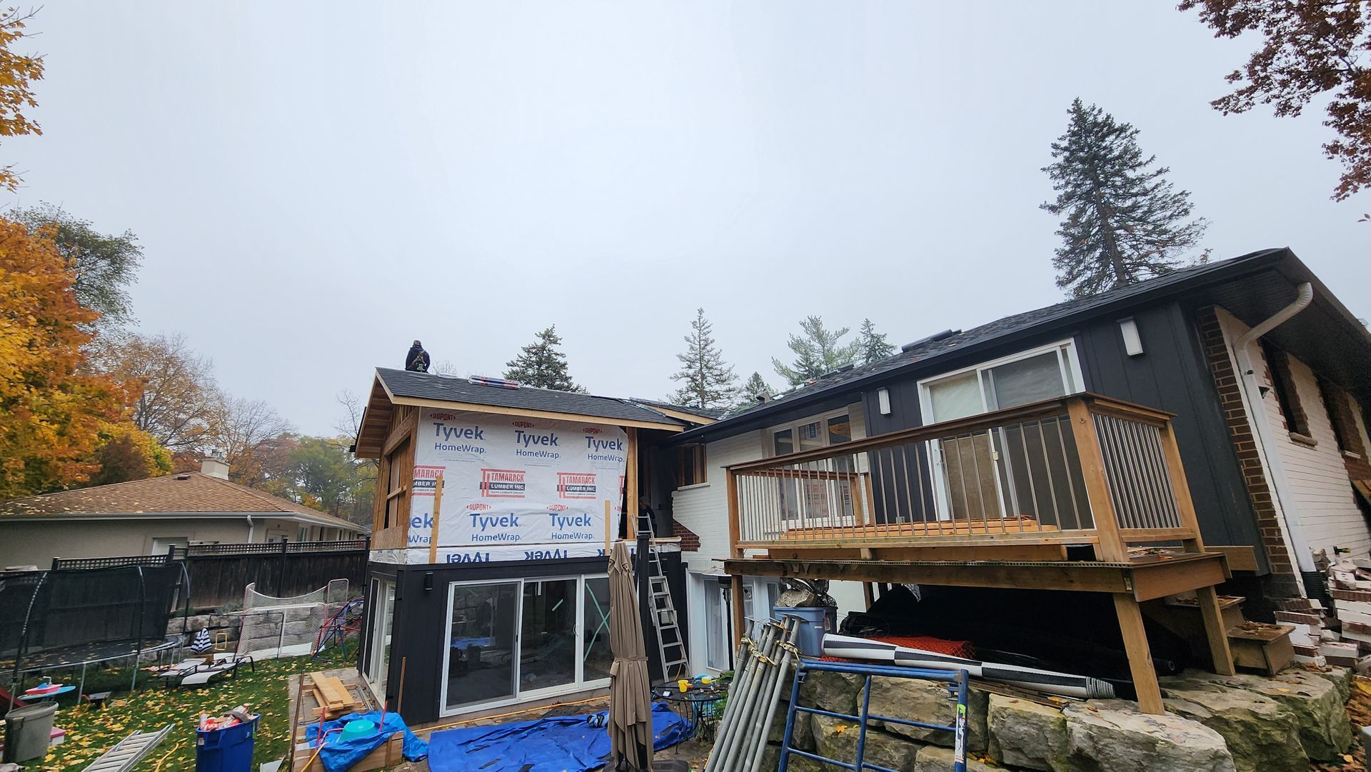 Partially built house with exposed framing and scaffolding beside a blue shed in an autumn yard
