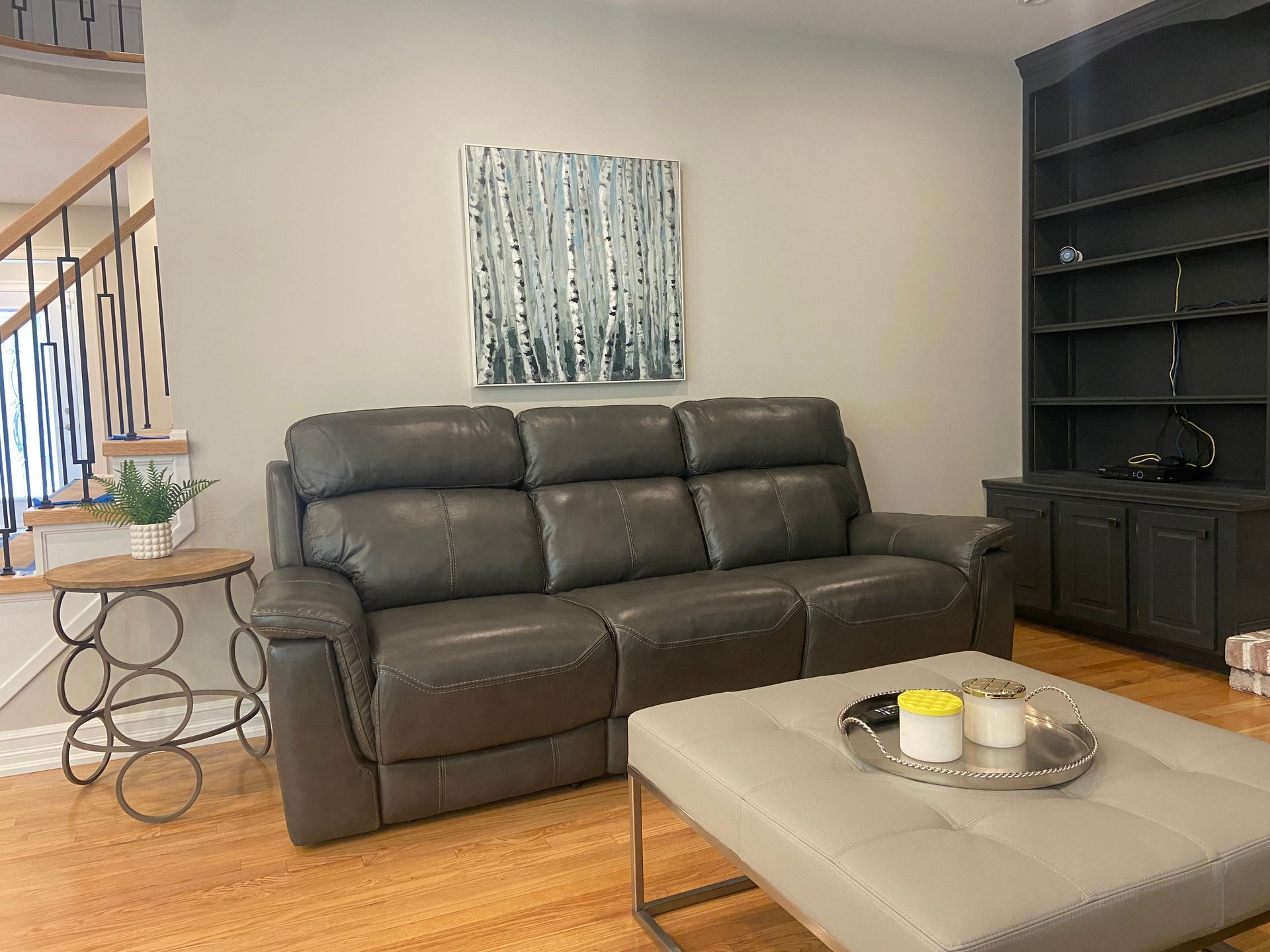 Living room with a dark leather sofa, beige ottoman, side table, and black shelving unit