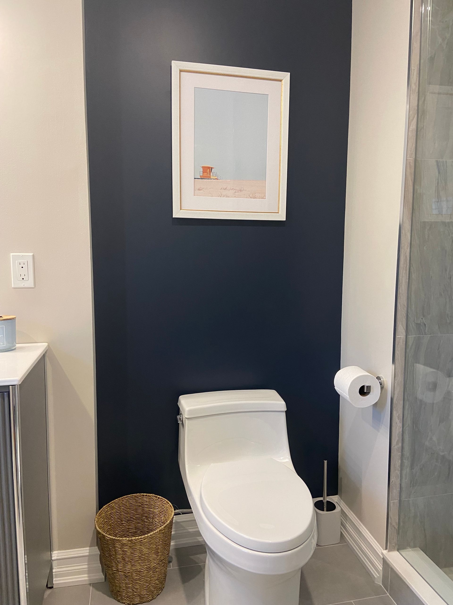 Small bathroom with navy accent wall, framed art, toilet, wicker basket, and glass shower door.