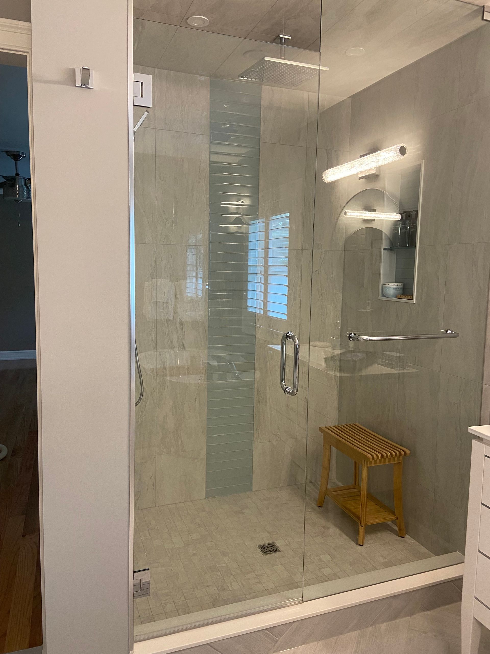 Glass shower enclosure in a tiled bathroom with a wooden stool and wall light.