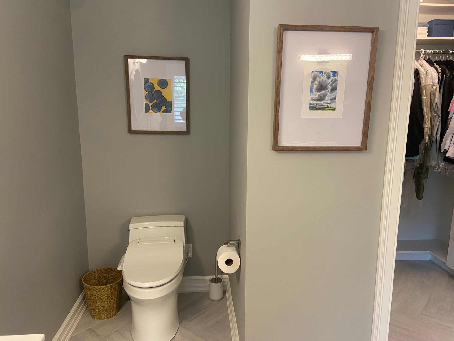 Small bathroom with a toilet, toilet paper holder, wicker basket, and two framed pictures on gray walls.