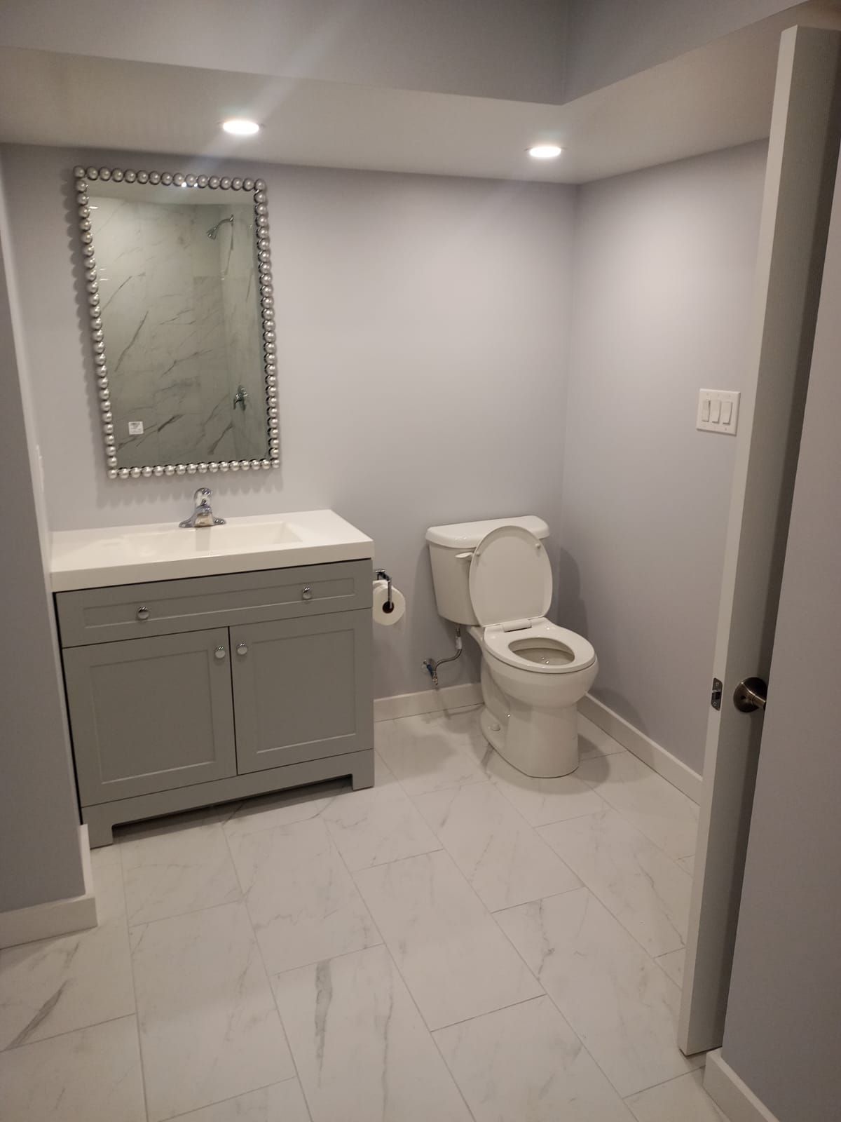 Small modern bathroom with gray vanity, mirror, toilet, and white tile floor