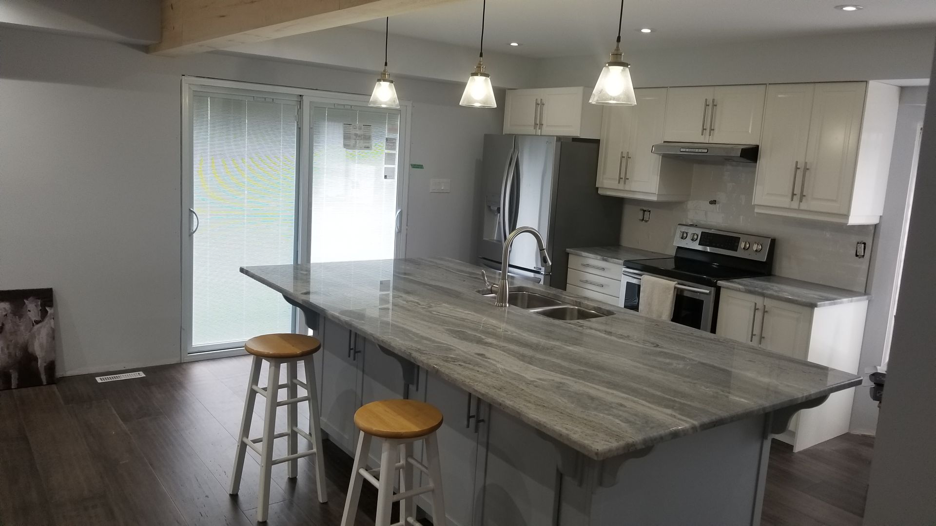 Bright kitchen with a large marble island, pendant lights, white cabinets, and stools by a window.