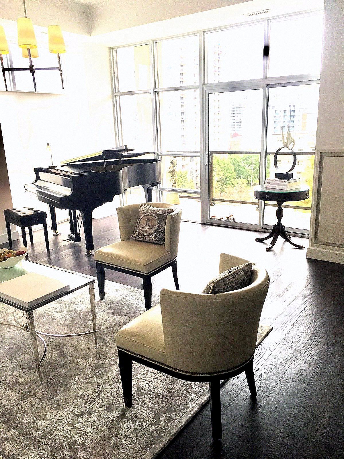 Bright living room with a grand piano, chairs, a rug, and large windows overlooking greenery.