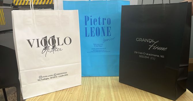 Tre borse della spesa di marca – bianca, blu e nera – appoggiate su una superficie di legno.