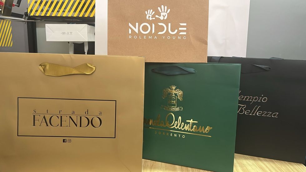 Su un tavolo sono esposte borse della spesa di diverse marche, nei colori oro, verde e nero.