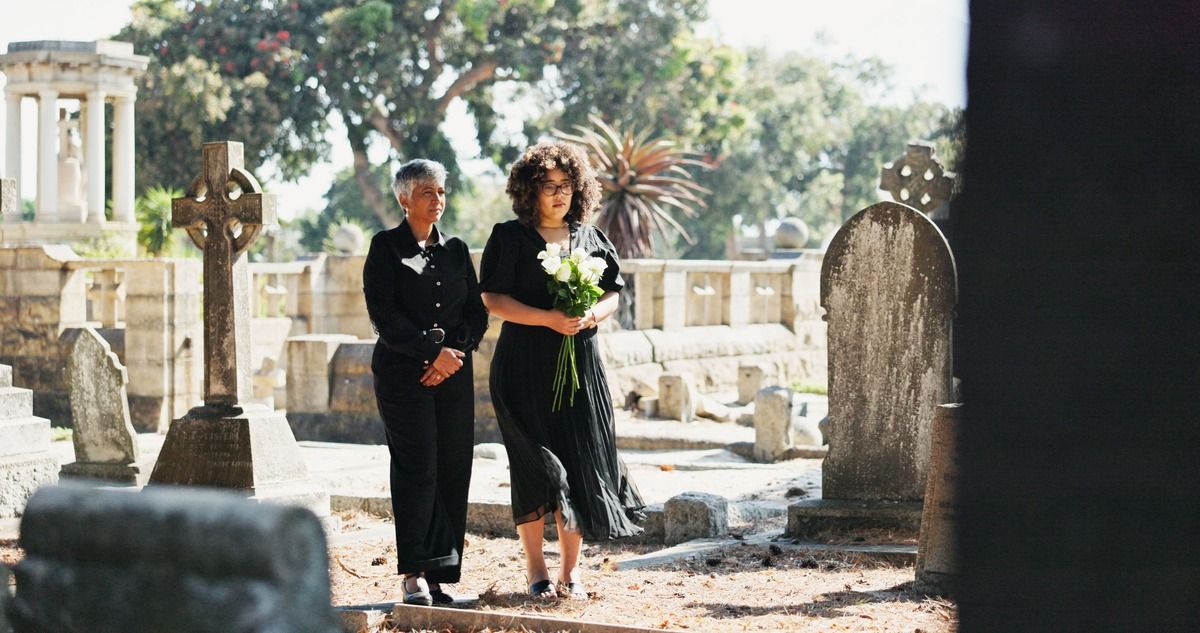 Two people in dark clothes walk through a sunlit cemetery among gravestones.